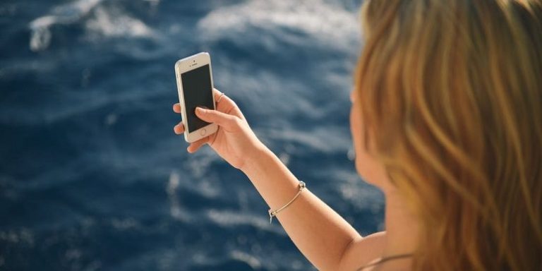 A girl holds her phone to take a selfie by the ocean and is likely not thinking about the link between social media and eating disorders