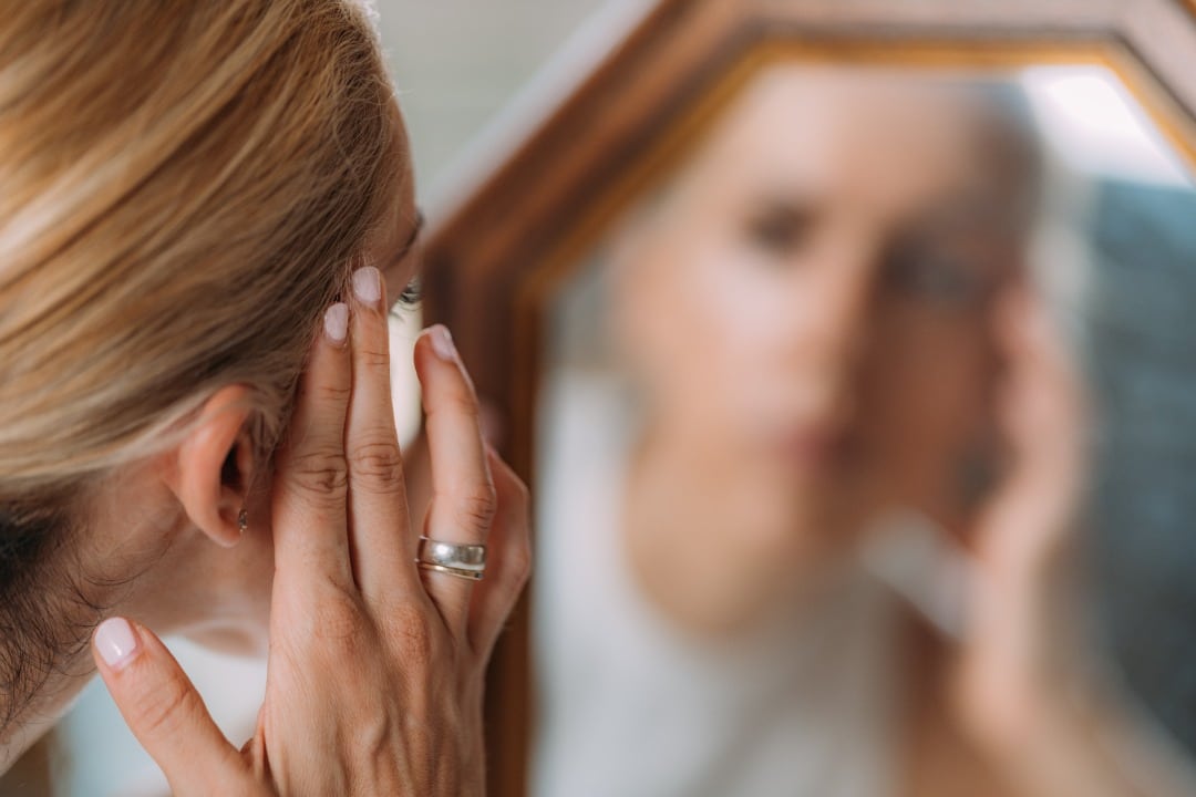 Woman looking at her reflection in a mirror, and pulling her face tighter with her hands.