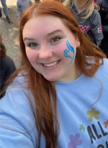 A smiling selfie of Emma. She has red hair, and has her face painted on the left side with a blue NEDA symbol.