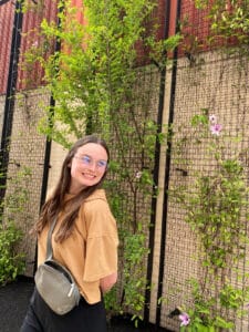 Lauren stands in front of a wall with plants. She's wearing glasses and smiling over her shoulder.